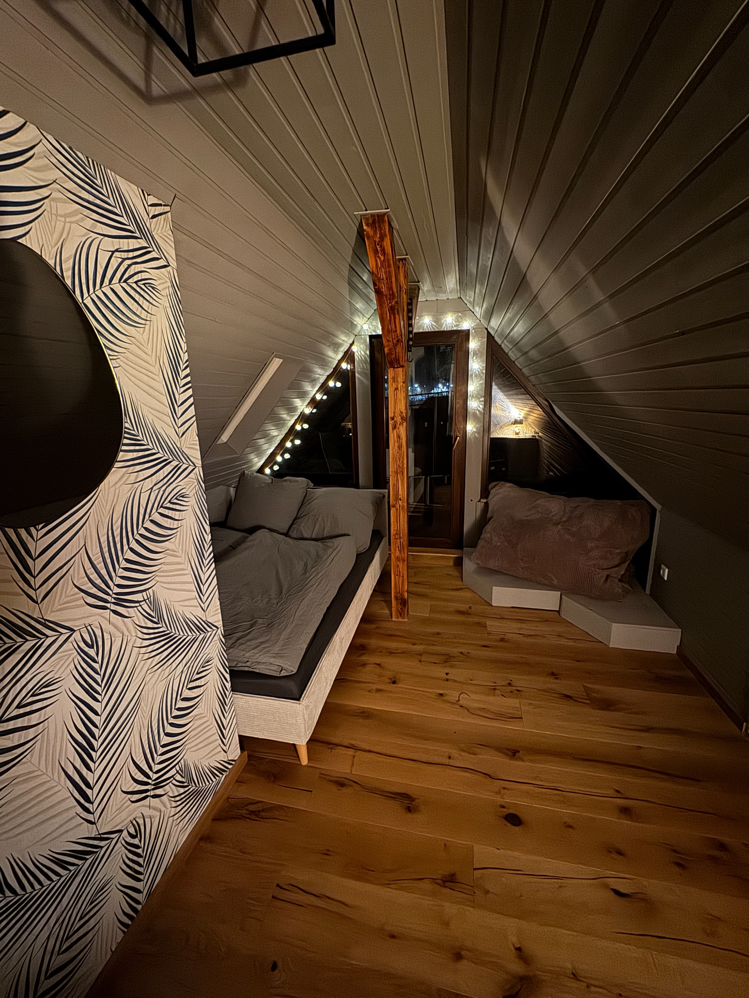Schlafzimmer unter der Dachschräge mit sichtbarem Holzbalken und warm beleuchtetem Fenster